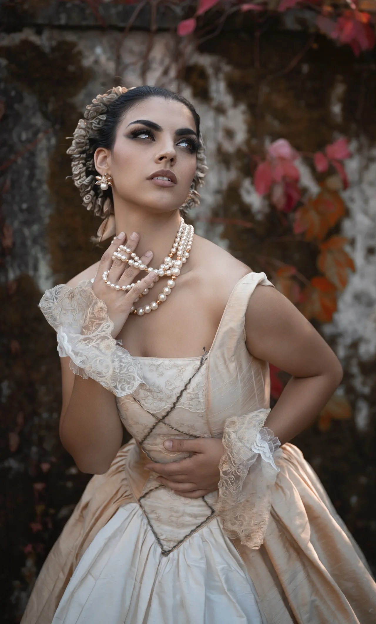 Woman in ivory elizabethan gown wearing 3 pearl necklaces. her Hand is underneath the necklaces holding them away from her neck